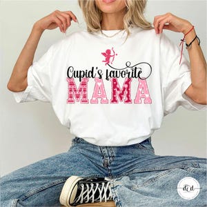 May include: White t-shirt with the text "Cupid's favourite MAMA" in pink and red lettering. A pink Cupid graphic is above the text. The shirt is worn with blue jeans and black trainers.
