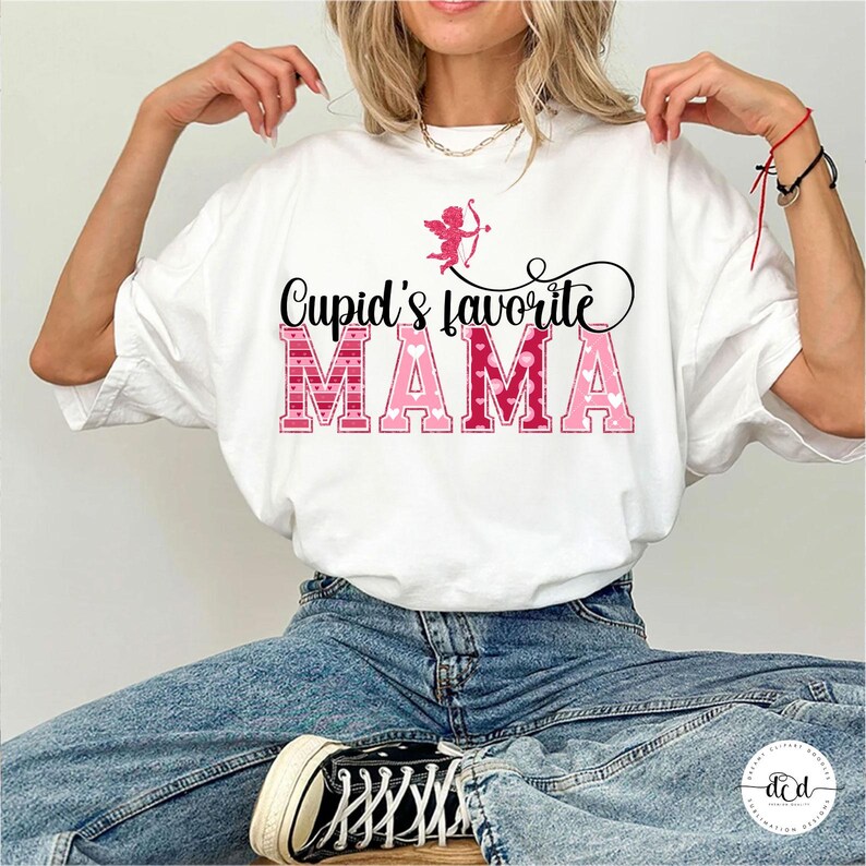 May include: White t-shirt with the text "Cupid's favourite MAMA" in pink and red lettering. A pink Cupid graphic is above the text. The shirt is worn with blue jeans and black trainers.