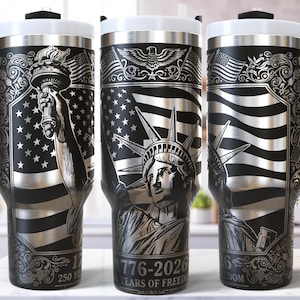 Op de afbeelding: Zwarte en zilveren reisbeker met een geëtst ontwerp van het Vrijheidsbeeld, Amerikaanse vlaggen en de tekst "1776-2026 Years of Freedom". De beker heeft een zwart handvat en een wit deksel.