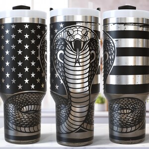 May include: Three stainless steel travel mugs with black handles and lids. The mugs feature unique designs: one with a star pattern, one with a cobra, and one with black and silver stripes. The mugs are designed for hot or cold beverages.