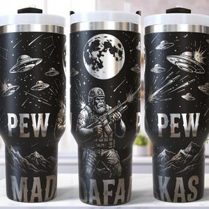 Pode incluir: Caneca de viagem preta com tampa e alça brancas, com um design de OVNIs, estrelas cadentes e uma figura semelhante a um soldado com uma arma sob uma lua cheia. O texto "PEW MADAFKAS" é exibido em letras prateadas.
