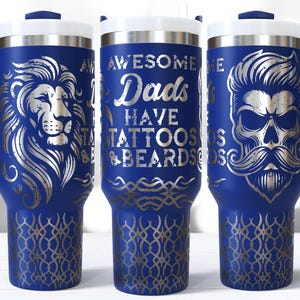 Puede incluir: Vaso térmico azul marino con tapa y asa blancas. El vaso presenta diseños plateados de un león, una calavera con bigote y el texto "Awesome Dads Have Tattoos & Beards". La parte inferior tiene un patrón decorativo.
