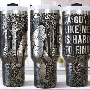 May include: A set of three stainless steel travel mugs with black handles and white lids. Each mug features a detailed silver engraving of a Bigfoot in a forest scene, with the text "A Guy Like Me Is Hard To Find".