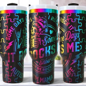 May include: Black tumbler with a handle, featuring a colorful puzzle piece pattern and the text "Some Days It Rocks Me." The design includes a skeleton graphic and the words "Autism Mama."