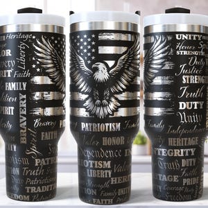 Op de afbeelding: Drie zwarte geïsoleerde bekers met witte deksels en handvatten. Elke beker heeft een ontwerp met een Amerikaanse vlag, een adelaar en patriottische woorden zoals "Vrijheid" en "Patriotisme".