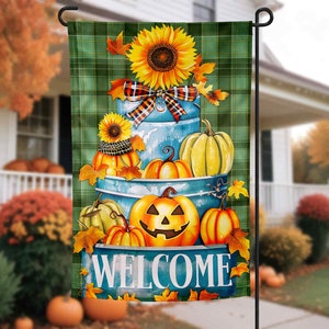 May include: A green and white plaid welcome garden flag with a stack of blue and white painted pumpkins, gourds, and a sunflower. The pumpkins are stacked on top of each other and the sunflower is on top of the stack. The flag says "Welcome" in white letters.
