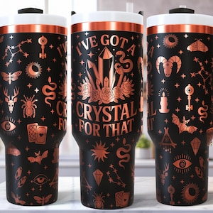 May include: Three black travel mugs with copper-colored accents and handles. Each mug features a different design with mystical symbols and the phrase "I've Got a Crystal For That." The mugs have white lids and are suitable for hot or cold beverages.