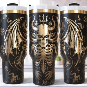 May include: Black travel tumbler with a gold skeleton, wings, and horns design. The tumbler has a black handle, a white top, and a gold rim. The design includes various symbols and a trident.