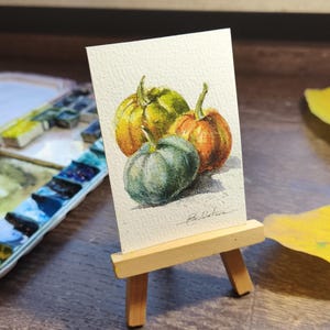 Può includere: Acquerello di tre zucche nei toni del verde, arancione e giallo. L'opera d'arte è esposta su un piccolo cavalletto di legno. Il dipinto è su carta bianca. Lo sfondo è sfocato.