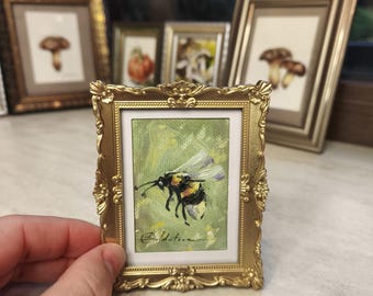 May include: A miniature painting of a bee in flight, set within a small, decorative gold frame. The artwork features a bee against a green and yellow backdrop. Other framed paintings are visible in the background, suggesting a collection.