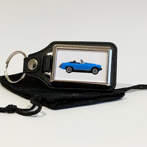 Op de afbeelding: Een blauwe cabriolet-sleutelhanger met een zwarte leren band en een zilveren metalen sleutelring. De sleutelhanger ligt op een zwarte stoffen tas met een trekkoord.