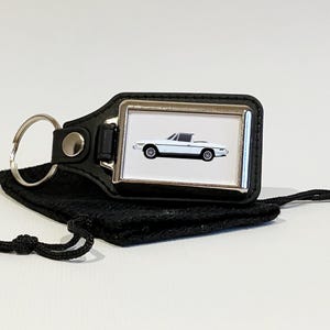 May include: A white leather keychain with a silver metal frame featuring a white convertible car. The keychain is attached to a black leather pouch with a drawstring.