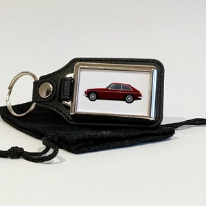Op de afbeelding: Een rode MG sportwagen sleutelhanger met een zwarte leren band en een zilveren metalen frame. De sleutelhanger is bevestigd aan een zwarte trekkoordtas.