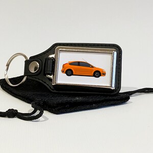 May include: A black leather keyring with a silver metal frame featuring an orange Ford Focus car. The keyring comes with a black drawstring bag.