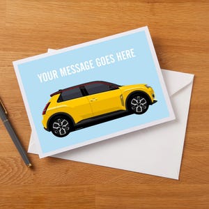 May include: Greeting card featuring a yellow car illustration on a light blue background. The card reads "YOUR MESSAGE GOES HERE" in white text. A pen rests on a wooden surface next to the card and white envelope.