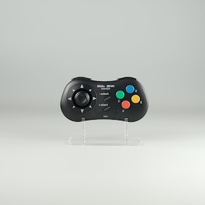 Puede incluir: Un mando de videojuego negro con un pad direccional circular, cuatro botones de colores, un botón de inicio y un botón de selección. El mando está etiquetado como "SNK NEOGEO" y está en un soporte transparente.