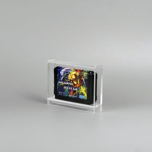 Puede incluir: Un cartucho de videojuego Sega Genesis negro con la etiqueta "Shadow Dancer" se muestra dentro de una caja de acrílico transparente. El cartucho presenta ilustraciones coloridas de un guerrero. La caja es rectangular y transparente.