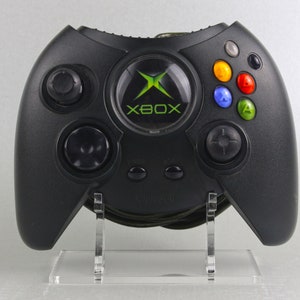 Könnte beinhalten: Ein schwarzer Xbox-Controller mit grünen und weißen Akzenten. Der Controller steht auf einem klaren Acryl-Ständer. Der Controller hat einen grünen X-Knopf mit dem Wort "Xbox" darauf.