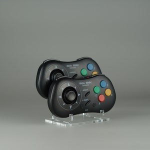 Puede incluir: Dos mandos de videojuegos negros con un joystick y botones. Los mandos están en un soporte de acrílico transparente. Los mandos tienen el texto "NEO GEO" y "SNK" en ellos.