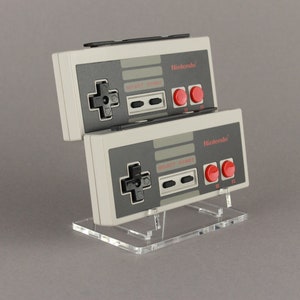 Könnte beinhalten: Zwei graue Nintendo Entertainment System-Controller mit roten Knöpfen werden auf einem durchsichtigen Acryl-Ständer präsentiert. Die Controller sind übereinander gestapelt.