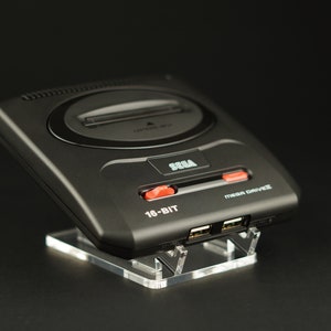 Op de afbeelding: Een zwarte Sega Genesis 16-bit Mega Drive console met rode knoppen en een transparante acryl standaard.
