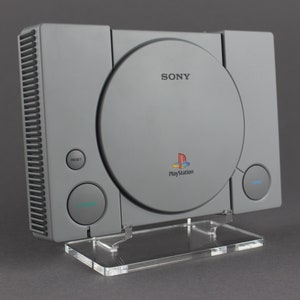 May include: A gray Sony PlayStation console with a clear plastic stand. The console has a disc drive on the front and the PlayStation logo is visible.