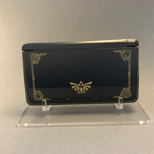 May include: A black Nintendo 3DS XL console with a gold Triforce design and decorative border. The console is on a clear acrylic stand.