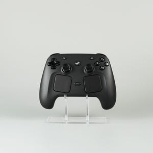 May include: A black Steam Controller game pad featuring a directional pad, two analogue sticks, four action buttons labelled A, B, X, and Y, two large touchpads, and a central Steam logo button, all mounted on a clear acrylic display stand.