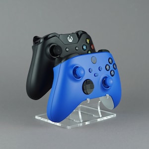 Könnte beinhalten: Zwei kabellose Gamecontroller, einer schwarz und einer blau, werden auf einem transparenten Acrylständer präsentiert. Der blaue Controller befindet sich im Vordergrund und zeigt seine Tasten und Joysticks. Der schwarze Controller befindet sich dahinter, mit dem Xbox-Logo.