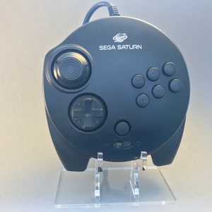 May include: A black Sega Saturn controller with a clear plastic stand. The controller has a circular design with a directional pad, six buttons, and a large, round button in the center. The text "SEGA SATURN" is printed on the front of the controller.
