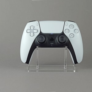 Könnte beinhalten: Ein weißer PlayStation 5 DualSense Wireless-Controller mit schwarzen Tasten, der auf einem transparenten Acryl-Ständer präsentiert wird. Der Controller befindet sich auf dem Ständer und der Ständer steht auf einer grauen Oberfläche.