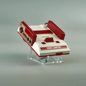 Op de afbeelding: Een witte en rode Nintendo Family Computer console met een rode controller bevestigd. De console staat op een transparante acryl standaard.