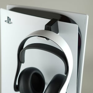 以下が含まれることがあります： 白と黒のゲーミングヘッドセットが、白いPlayStation 5コンソールに取り付けられた黒いホルダーに取り付けられています。コンソールにはPlayStationのロゴが側面にあります。ヘッドセットはゲーム用に設計されています。