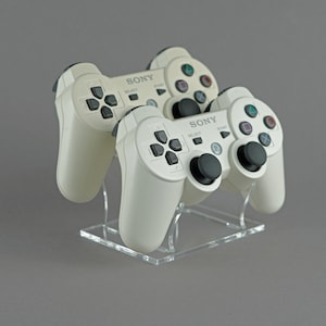 Op de afbeelding: Twee witte Sony PlayStation 3-controllers op een transparante acrylstandaard. De controllers hebben zwarte knoppen en een zwart PlayStation-logo.