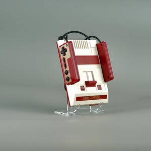 Könnte beinhalten: Eine weiße und rote Nintendo Entertainment System (NES) Konsole mit zwei angeschlossenen Controllern. Die Konsole steht auf einem klaren Acryl-Ständer. Die Konsole trägt den Schriftzug "NES" und "FAMILY COMPUTER".