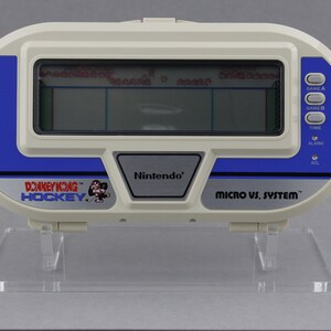 May include: A vintage Nintendo Micro vs. System Donkey Kong Hockey handheld game. The game has a white and blue plastic casing with a small LCD screen displaying the hockey game. The game has buttons for Game A, Game B, Time, Alarm, and ACL.