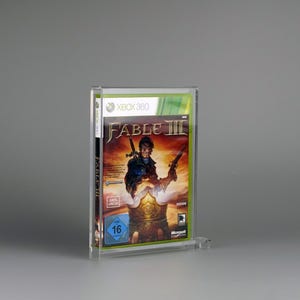 Könnte beinhalten: Eine transparente Acryl-Vitrine mit dem Spiel Fable III für Xbox 360. Das Cover des Spiels zeigt einen Krieger mit Schwert und Pistole vor einem feurigen Hintergrund. Die Vitrine dient zum Schutz und zur Präsentation des Spiels.