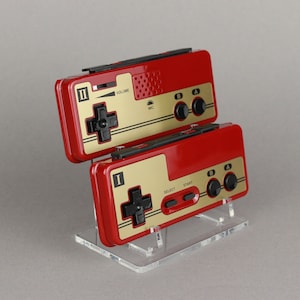 May include: Two red and gold handheld video game consoles with black buttons and a directional pad. The consoles are on a clear acrylic stand.
