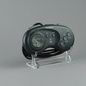 Könnte beinhalten: Ein schwarzer Sega Genesis-Controller mit einer grauen Frontplatte und einem schwarzen Kabel. Der Controller hat einen D-Pad, sechs Tasten und einen Startknopf. Der Controller steht auf einem durchsichtigen Plastikständer.