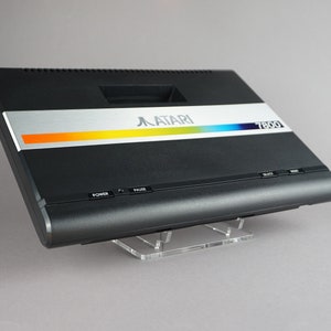 May include: A black Atari 7800 video game console with a silver and rainbow stripe on the front. The console has a power button, a pause button, and a reset button.