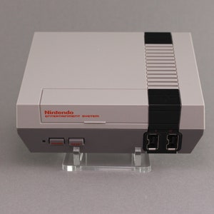Op de afbeelding: Een witte Nintendo Entertainment System console met een zwarte bovenkant en controllerpoorten. De console staat op een transparante acryl standaard.