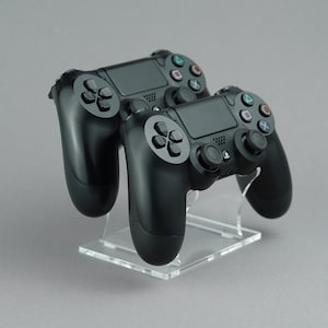 Puede incluir: Dos mandos negros de PlayStation 4 en un soporte de acrílico transparente. Los mandos están en posición vertical con la parte delantera hacia el espectador.