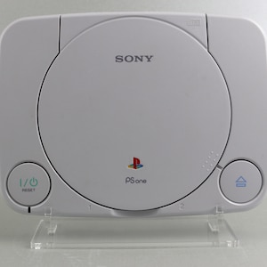 May include: A white Sony PlayStation One console with a black PlayStation logo and the text "PS one" on the front. The console has a disc drive slot and two buttons on the front. The console is sitting on a clear plastic stand.