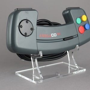 以下が含まれることがあります： 黒いジョイスティックと4つの色のボタンが付いた灰色のAmiga CD32コントローラー。コントローラーは透明なアクリルスタンドの上に置かれています。