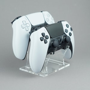 May include: Two PlayStation 5 DualSense controllers, one white and one black, are displayed on a clear acrylic stand. The controllers are positioned side-by-side, with the white controller on the left and the black controller on the right.