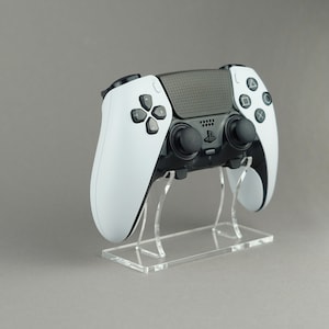 Op de afbeelding: Een witte en zwarte PlayStation 5-controller op een transparante acrylstandaard. De controller heeft een getextureerde zwarte bovenkant en witte handgrepen.