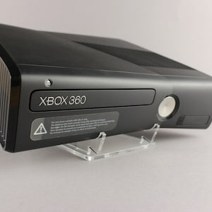 Puede incluir: Consola Xbox 360 negra con el texto "XBOX 360" en la parte delantera. La consola está sobre un soporte de acrílico transparente. La bandeja de disco y el botón de encendido son visibles. También hay una pegatina de advertencia.