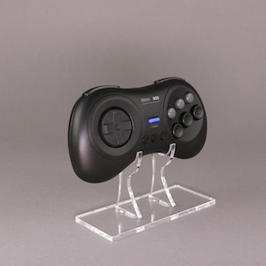 Könnte beinhalten: Ein schwarzer Sega Genesis-Controller mit 6 Tasten auf einem transparenten Acryl-Ständer. Der Controller verfügt über ein Steuerkreuz, sechs Tasten und einen Startknopf.