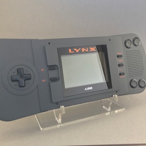 Könnte beinhalten: Eine schwarze Atari Lynx Handheld-Videospielkonsole mit einem grauen Bildschirm und orangefarbenem Text, der "LYNX" und "ATARI" lautet. Die Konsole steht auf einem durchsichtigen Acryl-Ständer.