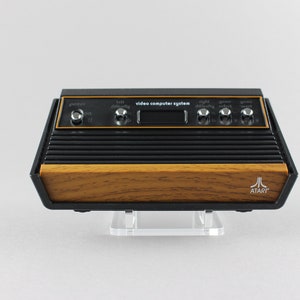 Può includere: Una console per videogiochi Atari vintage con un pannello frontale in legno e una parte superiore nera. La console ha un piccolo display digitale e diversi pulsanti per regolare le impostazioni. Il logo Atari è visibile sul pannello frontale.